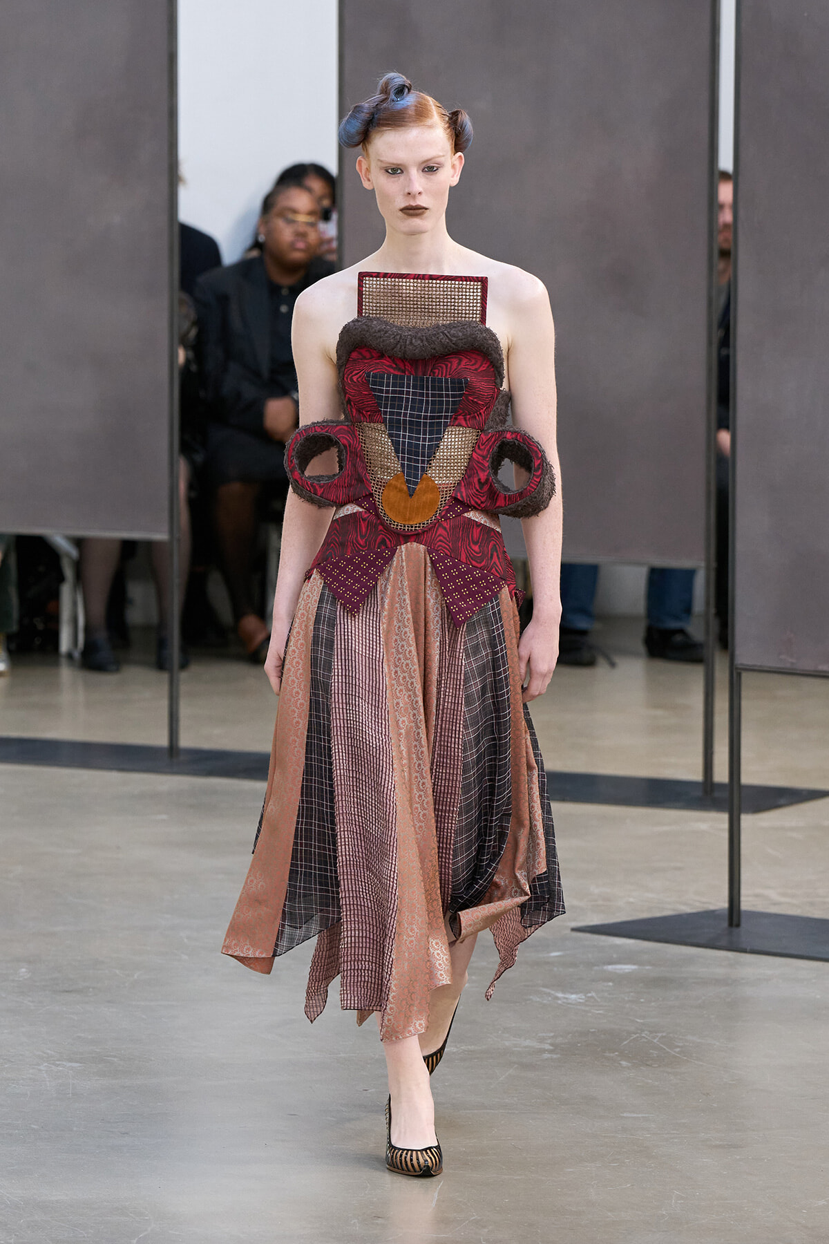 Avant-garde dress on a model featuring geometric, patterned panels in rust, burgundy, and black with sculptural shoulder details.