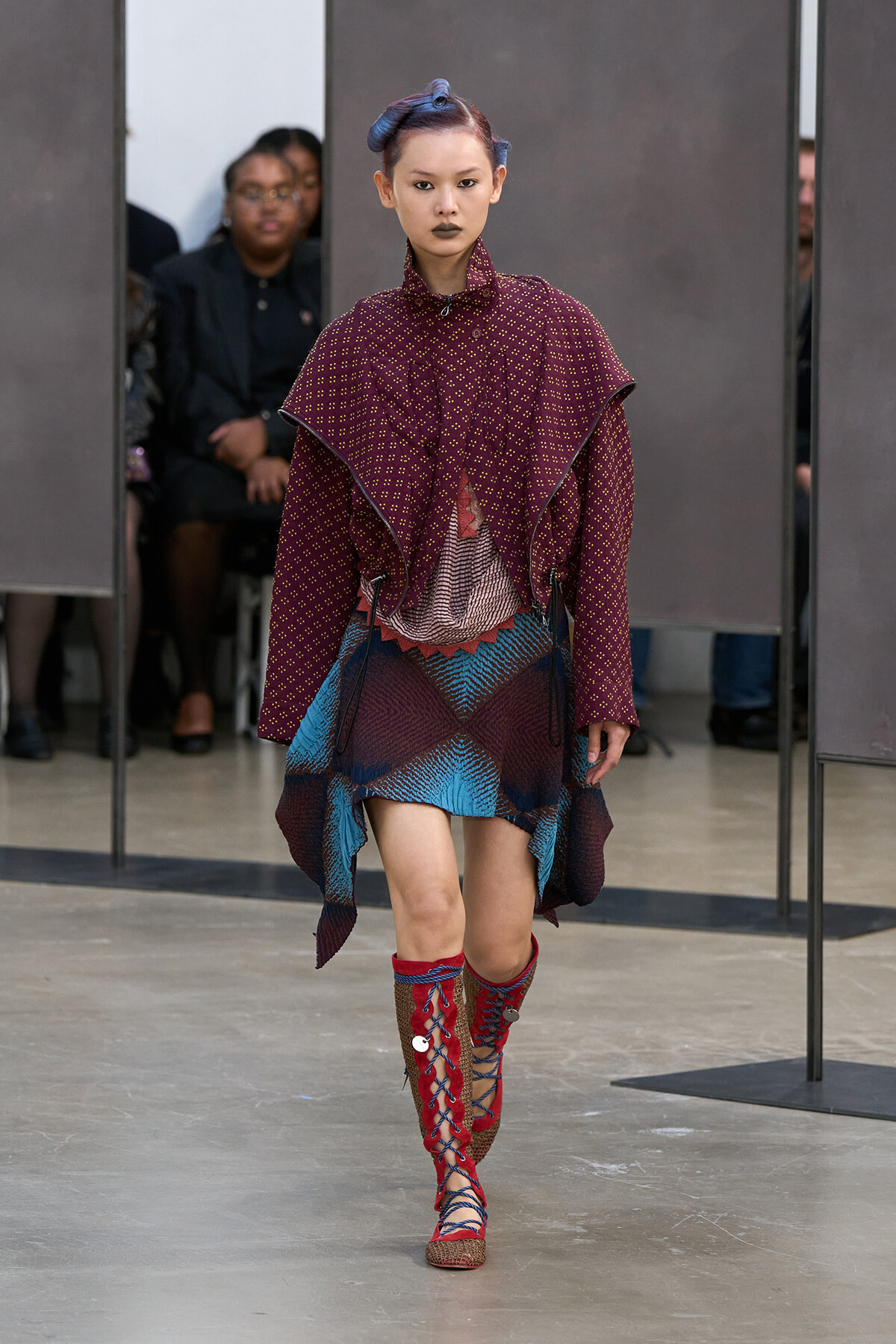 Model walks a runway wearing a burgundy patterned cape over a layered dress with a blue geometric skirt and red lace-up tall boots.