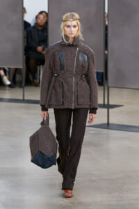 Model walking the runway in a textured brown coat with leather accents, matching pants, and a teardrop headpiece, carrying a brown textured bag with a blue bottom.