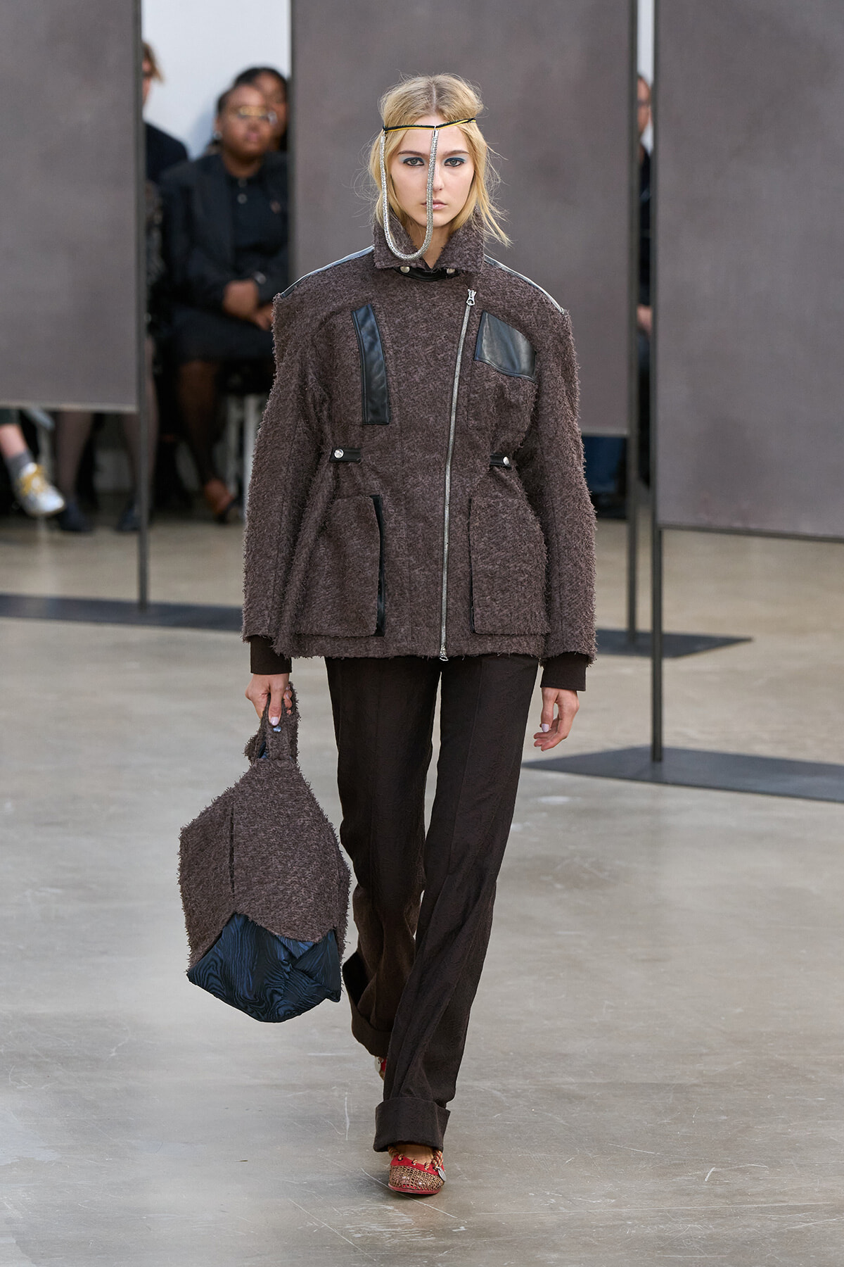 Model walking the runway in a textured brown coat with leather accents, matching pants, and a teardrop headpiece, carrying a brown textured bag with a blue bottom.