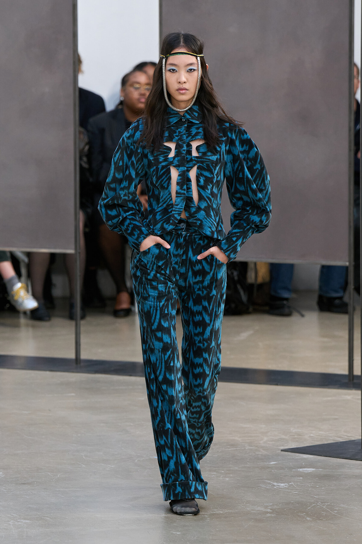 Model walking the runway in a teal-blue abstract print suit with cutout blouse and hands in pockets, wearing a thin headband accessory.
