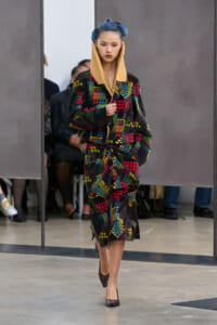 Model walking a runway in a bold, color-blocked patchwork coat and skirt with a cream shearling collar and blue hair.