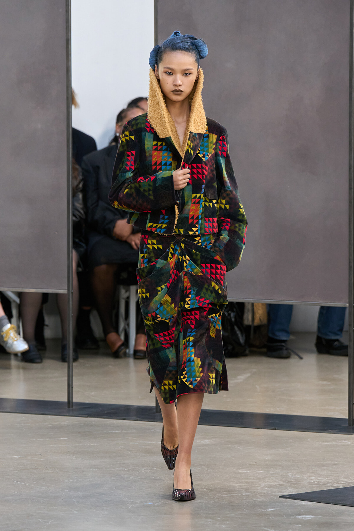 Model walking a runway in a bold, color-blocked patchwork coat and skirt with a cream shearling collar and blue hair.