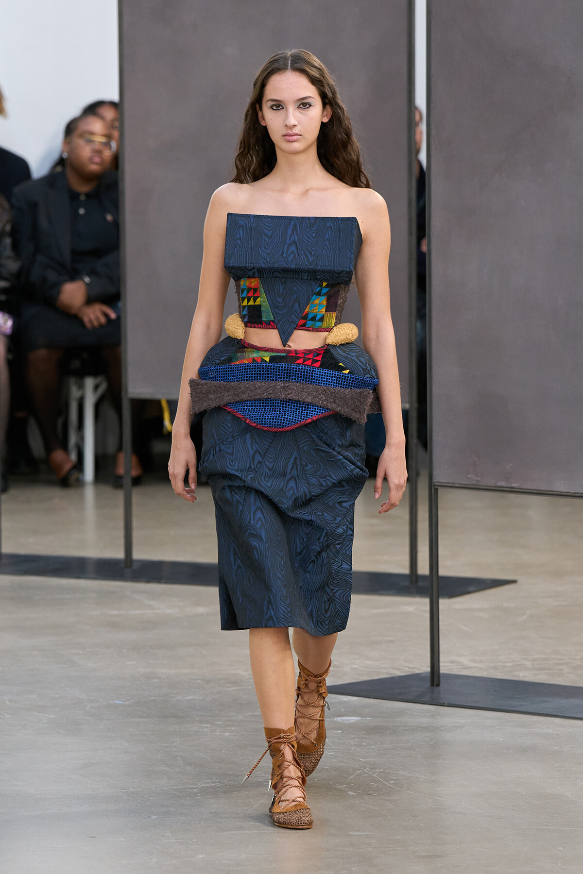 Model walks a fashion show wearing a dark blue textured strapless dress with a multicolored geometric waist panel and tan lace-up sandals.