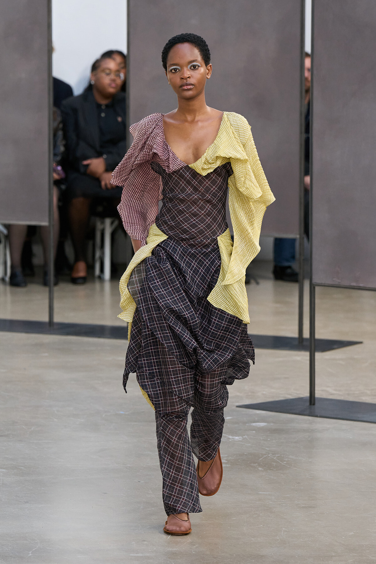 Model walking a runway in a brown plaid dress with yellow ruffled overlay.