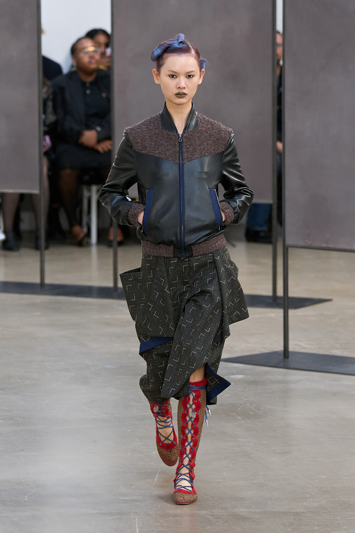 Model walking a fashion runway in a dark leather jacket with textured shoulders, layered gray patterned skirt, and red lace-up sandals. Audience blur in background.