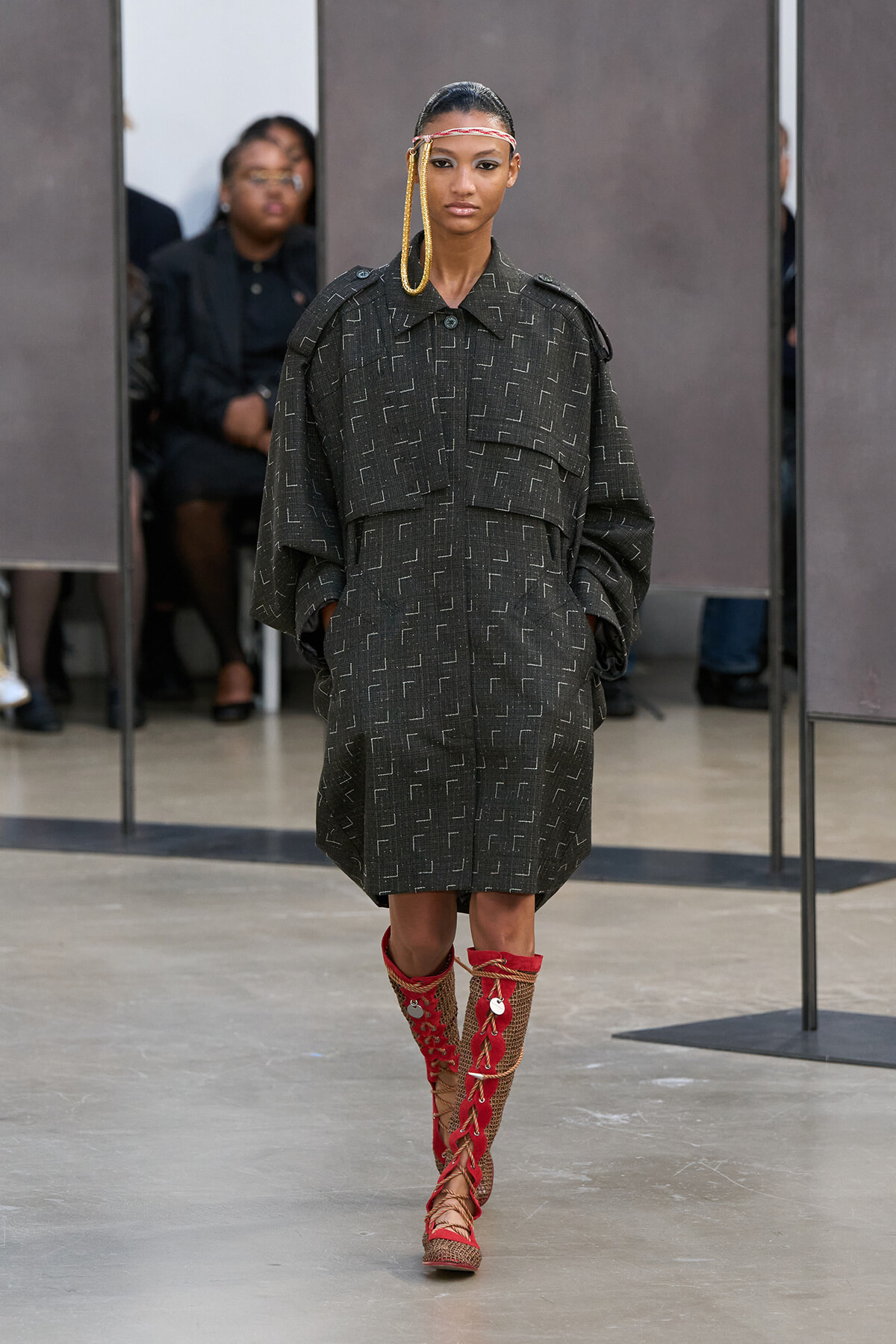 Model on a runway wearing an oversized charcoal coat with a subtle geometric pattern, paired with red lace-up knee-high boots.