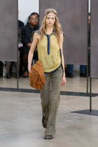 Model walking the runway in a yellow sleeveless blouse with blue trim and striped wide-leg pants, carrying a brown woven bag