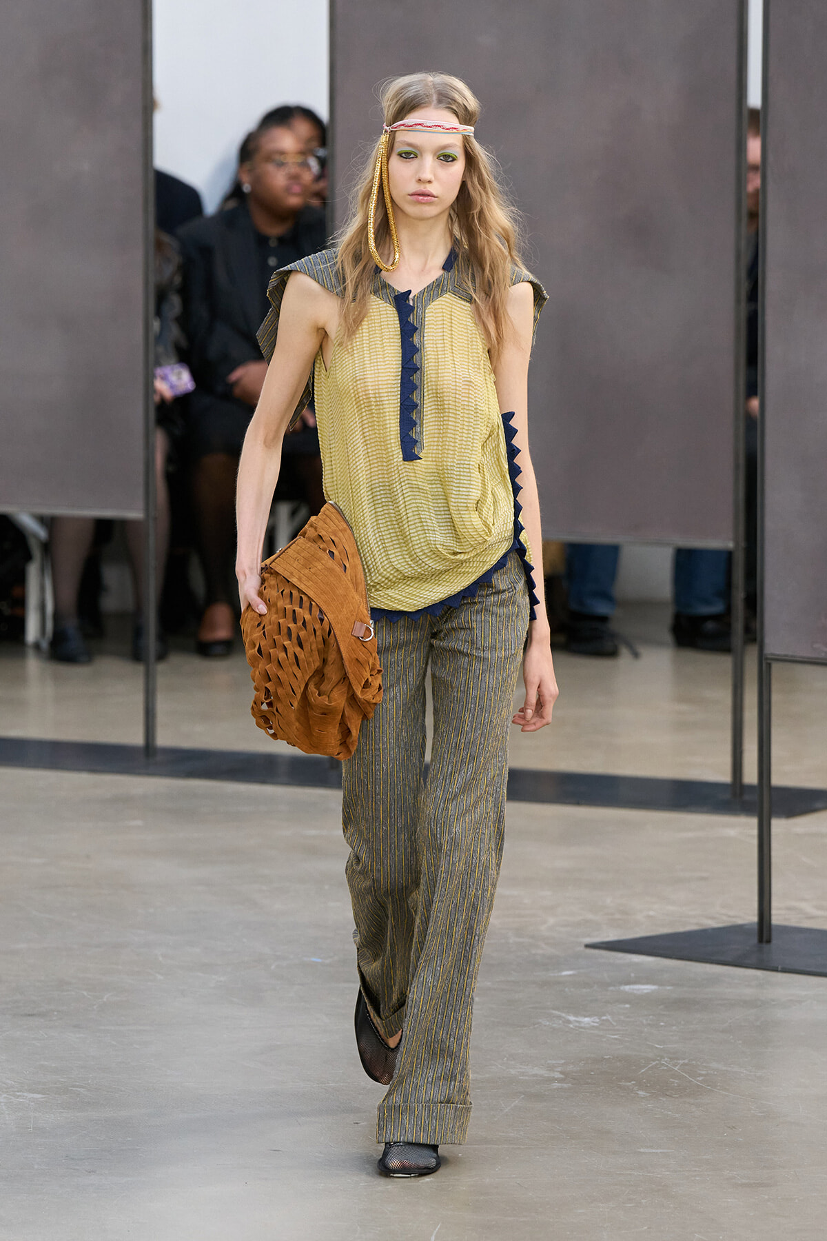 Model walking the runway in a yellow sleeveless blouse with blue trim and striped wide-leg pants, carrying a brown woven bag