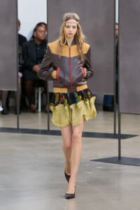Female model walks the runway in a brown leather jacket with yellow shearling, paired with a colorful, voluminous bubble skirt and headband.