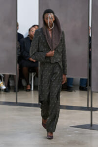 Model walks the runway in a dark gray patterned suit with a draped hood and a silver loop neck accessory.