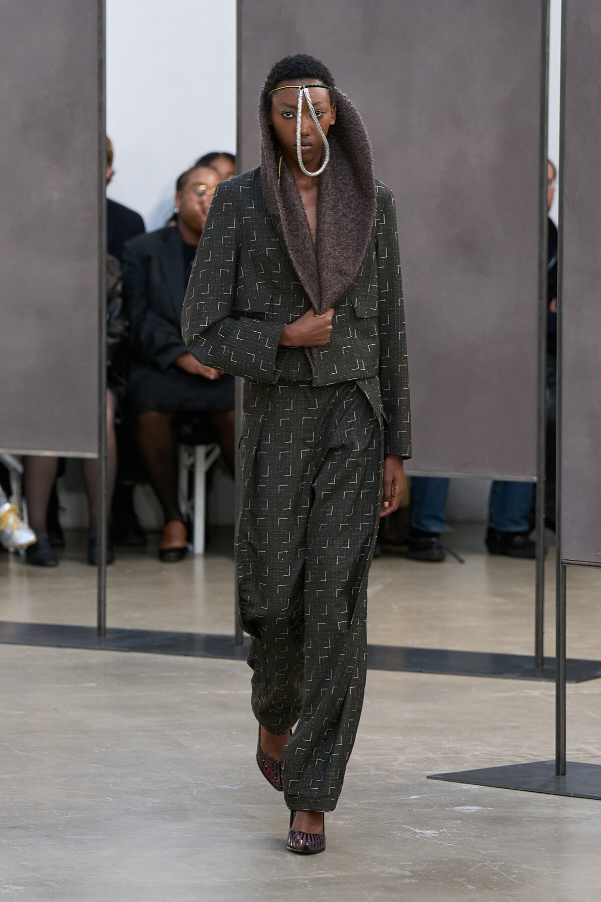 Model walks the runway in a dark gray patterned suit with a draped hood and a silver loop neck accessory.