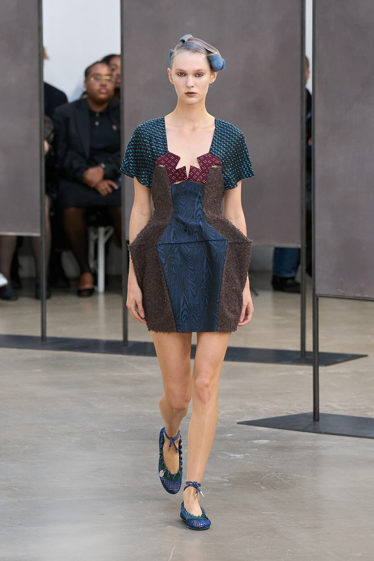 Model walks runway in an avant-garde navy dress with brown side panels and teal patterned top, paired with blue woven flats and hair rollers visible.