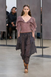 Model on a runway wearing a pink ruffled off-the-shoulder top with voluminous sleeves and loose plaid harem pants, tan lace-up boots, and a gold facial chain accessory.