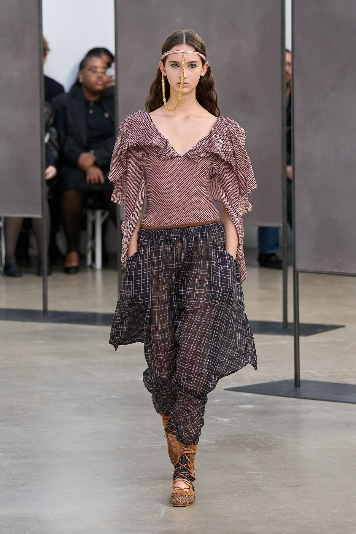 Model on a runway wearing a pink ruffled off-the-shoulder top with voluminous sleeves and loose plaid harem pants, tan lace-up boots, and a gold facial chain accessory.