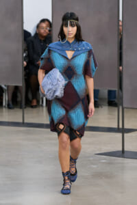 Model walks a runway in a blue geometric-patterned knit dress with a blue cape-like collar and a silver headpiece stilling over her forehead.