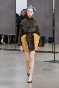 Model walking runway in a black patterned jacket with tan textured panels and blue hair, wearing high heels.