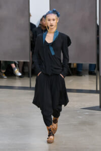 Model walking runway in loose black ensemble with blue patterned scarf, bold gold jewelry, and blue hair tied up, open stance with hands in pockets.
