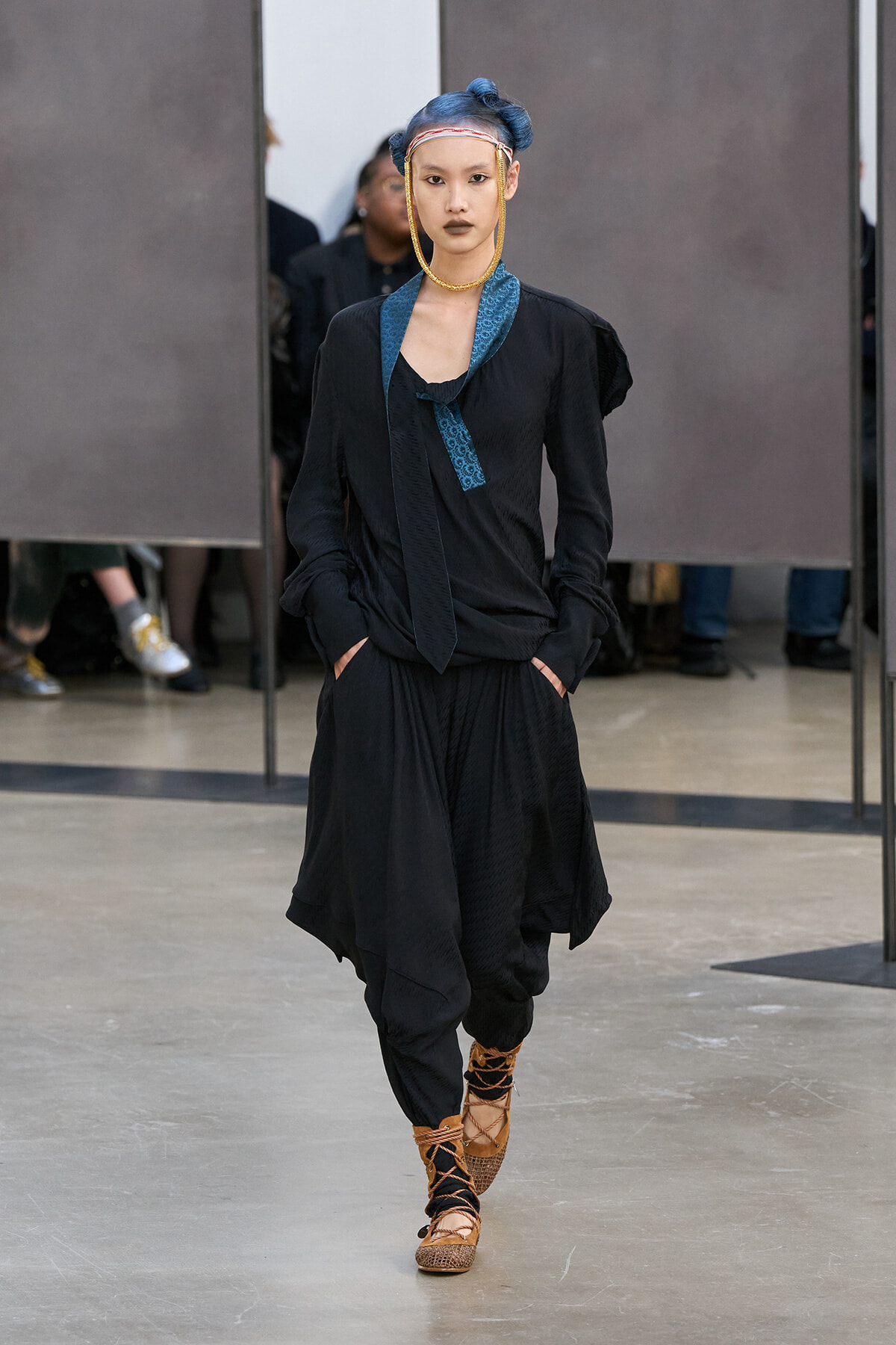 Model walking runway in loose black ensemble with blue patterned scarf, bold gold jewelry, and blue hair tied up, open stance with hands in pockets.