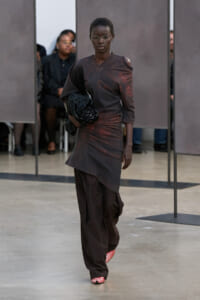 Model walking the runway in a textured dark brown asymmetric top and matching wide-leg pants, carrying a black woven clutch and wearing red-and-white striped shoes.