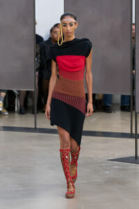 Female model on a runway wearing a color-block knit dress in black, red, brown, and navy with red lace-up knee-high boots.