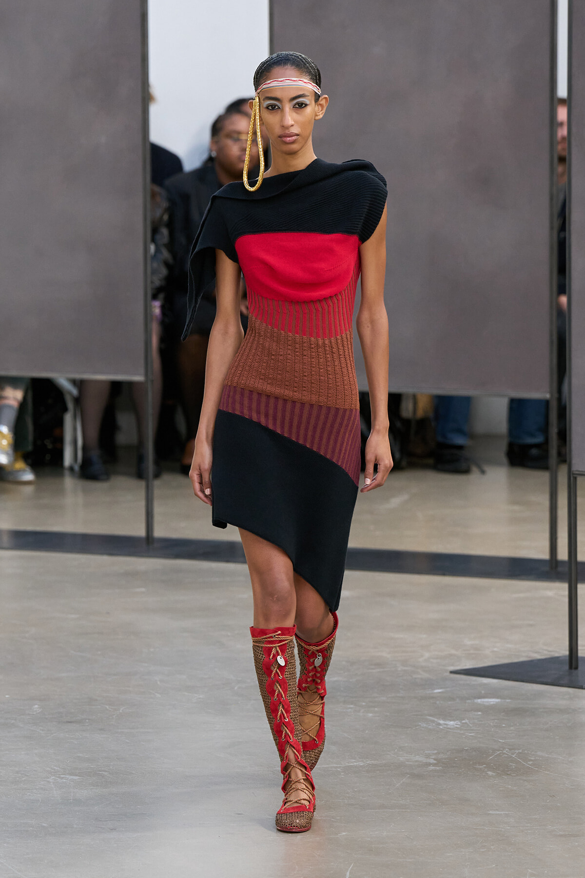 Female model on a runway wearing a color-block knit dress in black, red, brown, and navy with red lace-up knee-high boots.