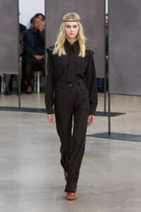 Female model walks the runway in a dark brown jumpsuit with black vest panels, accessorized with a headband and silver necklace.