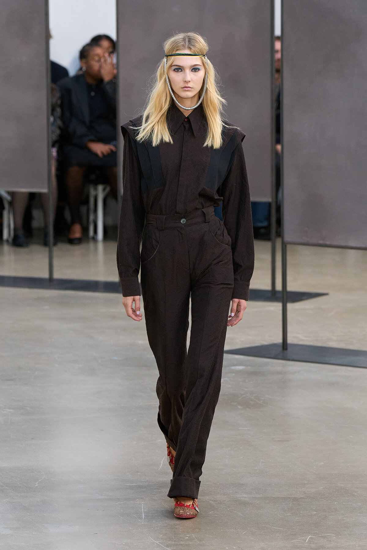 Female model walks the runway in a dark brown jumpsuit with black vest panels, accessorized with a headband and silver necklace.