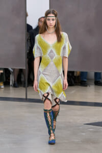 Model walking runway in a geometric lime-yellow and beige knit dress with wide sleeves; blue lace-up knee-high boots.