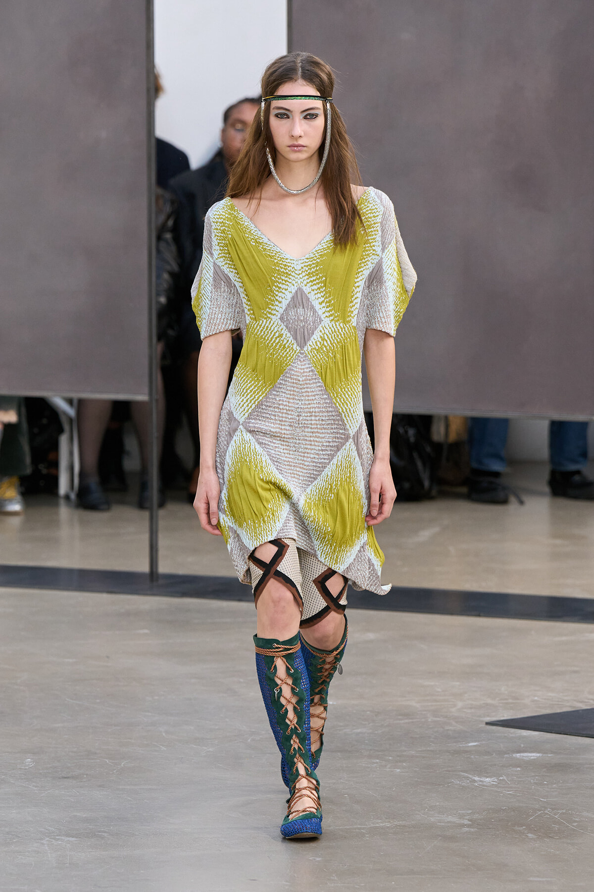 Model walking runway in a geometric lime-yellow and beige knit dress with wide sleeves; blue lace-up knee-high boots.