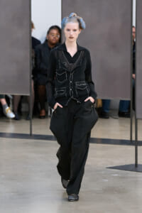 Model on a runway wearing a black stitched jacket and matching wide-leg pants; blue‑gray hair styled in two buns.