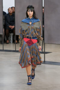 Model walks the runway in a blue-gray fringed dress with brown tufting, a red waist sash, and blue lace-up heels.