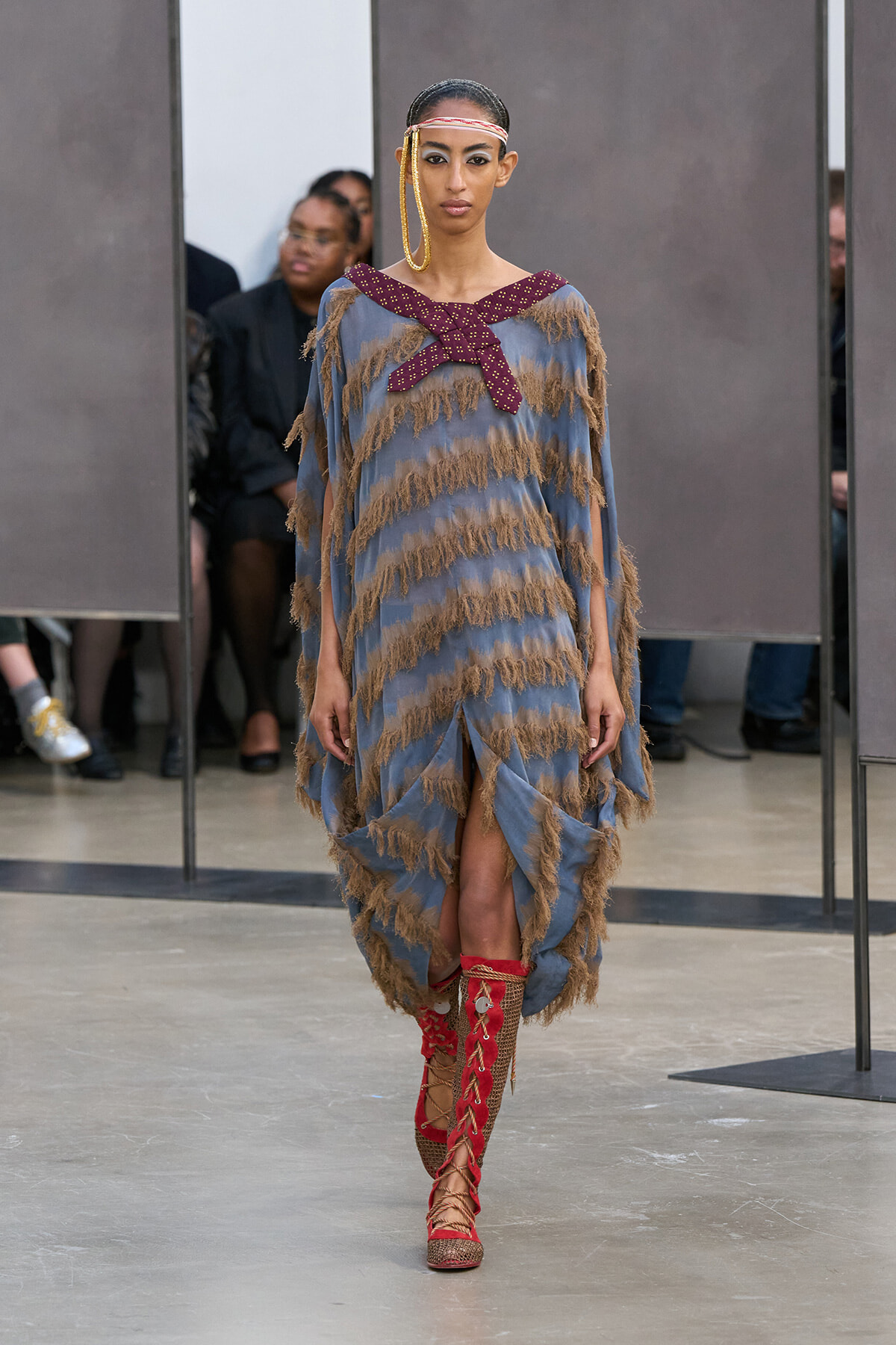 Model walking the runway in a blue fringe dress with tan fringe accents, paired with red lace-up boots and large gold hoop earrings.