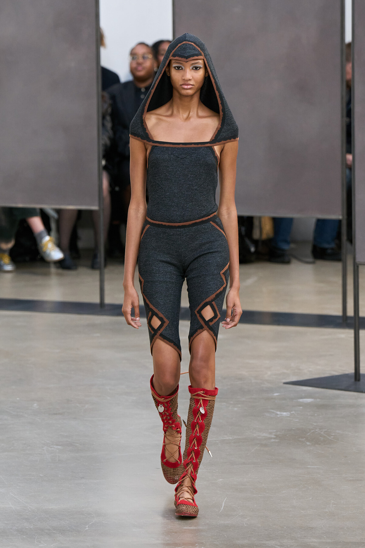 Model on a runway wearing a dark gray knit two-piece with orange trim and a hood, featuring geometric cut-outs and red lace-up boots.