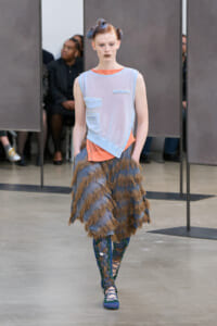 Model on a runway wearing a pale blue sleeveless top with pocket patches over an orange underlayer, paired with a blue-brown fringed skirt and blue lace-up boots.