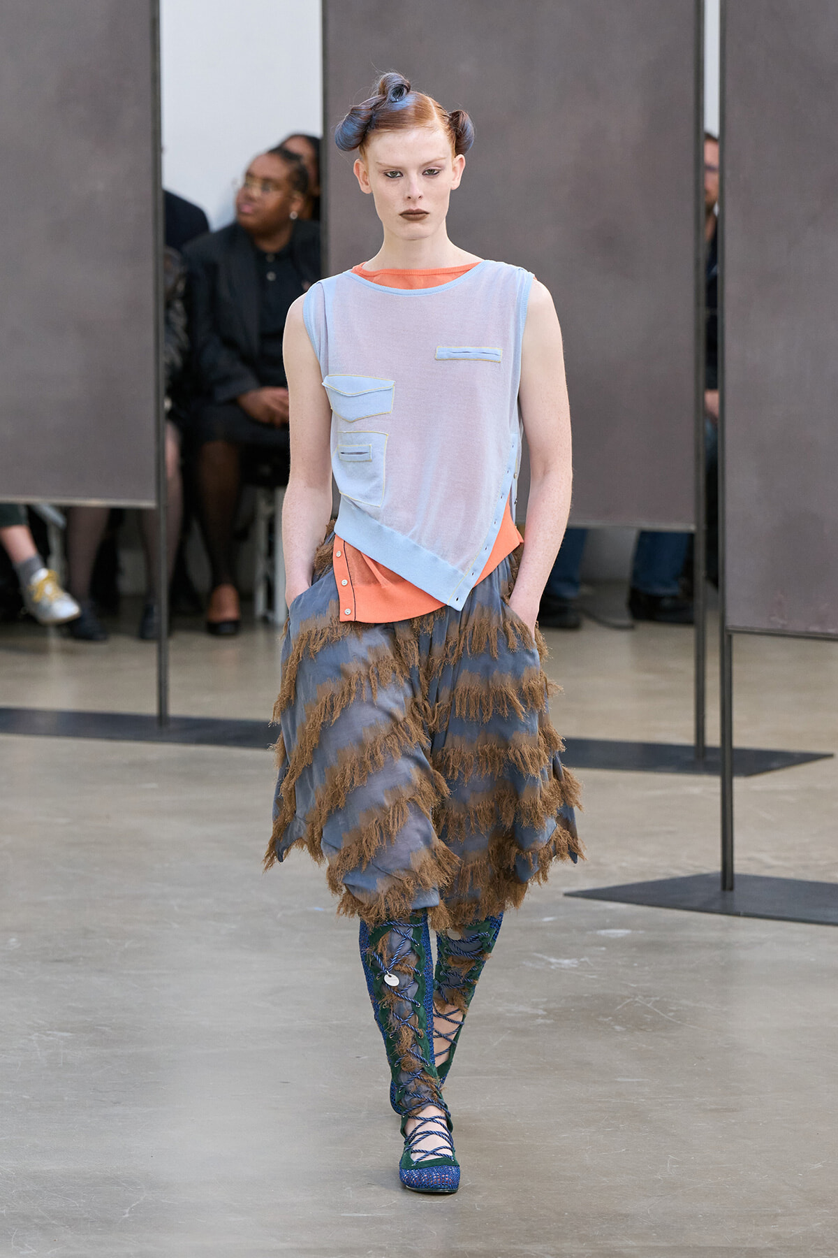 Model on a runway wearing a pale blue sleeveless top with pocket patches over an orange underlayer, paired with a blue-brown fringed skirt and blue lace-up boots.