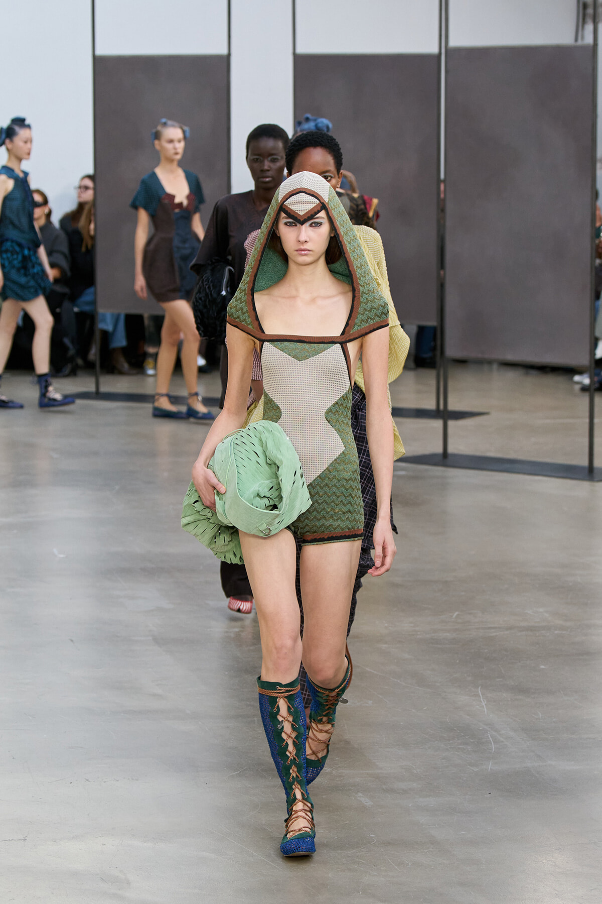 Model walks the runway in a green geometric hooded dress, holding a folded pale-green accessory at her side.