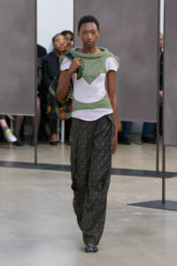 Model walks a fashion runway wearing a green textured top with white chevron panels and loose dark patterned pants, carrying a colorful bag