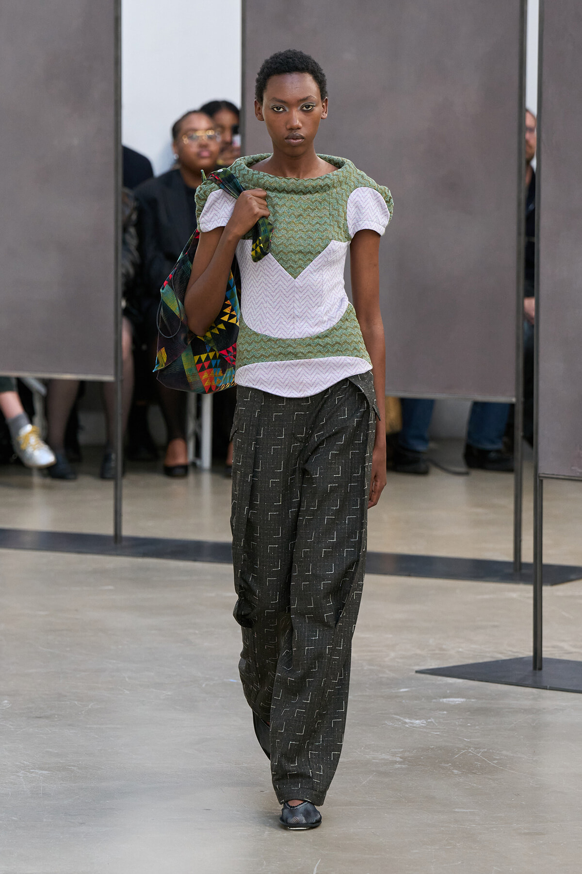 Model walks a fashion runway wearing a green textured top with white chevron panels and loose dark patterned pants, carrying a colorful bag