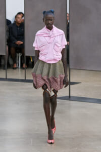 Model walking a fashion runway in a pink top with multiple pocket details and a layered striped skirt, wearing red-and-white striped heels.