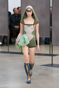 Model on a runway wearing a geometric green and beige hooded mini dress, holding a pale green wrap and blue lace-up boots.