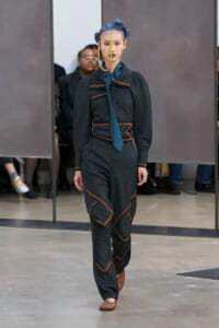 Female model walking runway in dark gray suit with copper piping, blue patterned tie, and blue braided hair with a gold chain headpiece