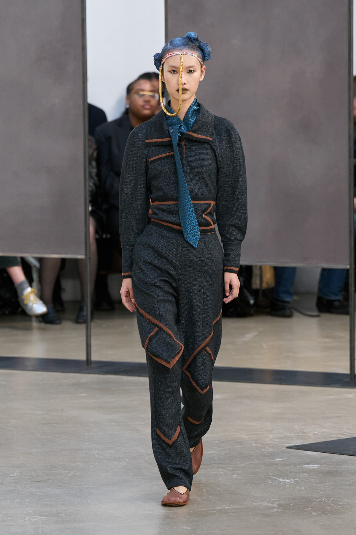 Female model walking runway in dark gray suit with copper piping, blue patterned tie, and blue braided hair with a gold chain headpiece