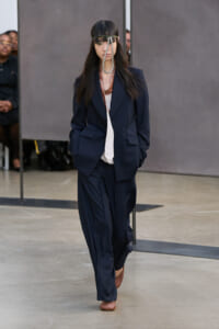 Model walking a fashion show in a navy oversized suit with wide-leg pants, white top, and brown shoes, accessorized with a metallic headband.