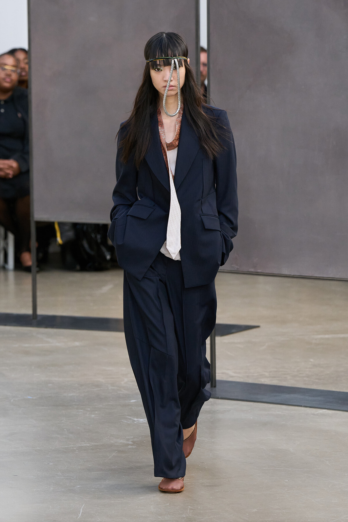 Model walking a fashion show in a navy oversized suit with wide-leg pants, white top, and brown shoes, accessorized with a metallic headband.