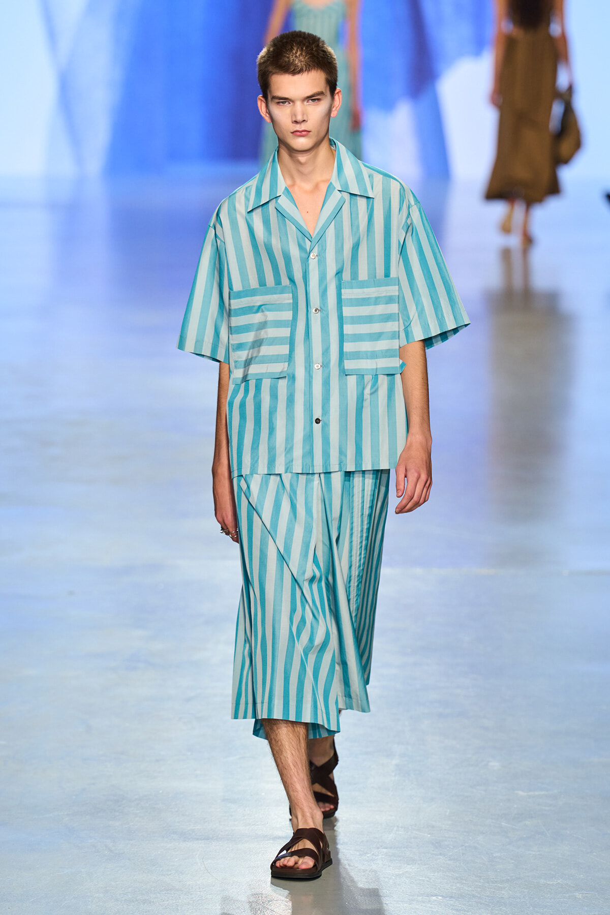 Male model walks the runway in a turquoise and light-blue striped short-sleeve shirt with matching striped pants and brown sandals in a fashion show.