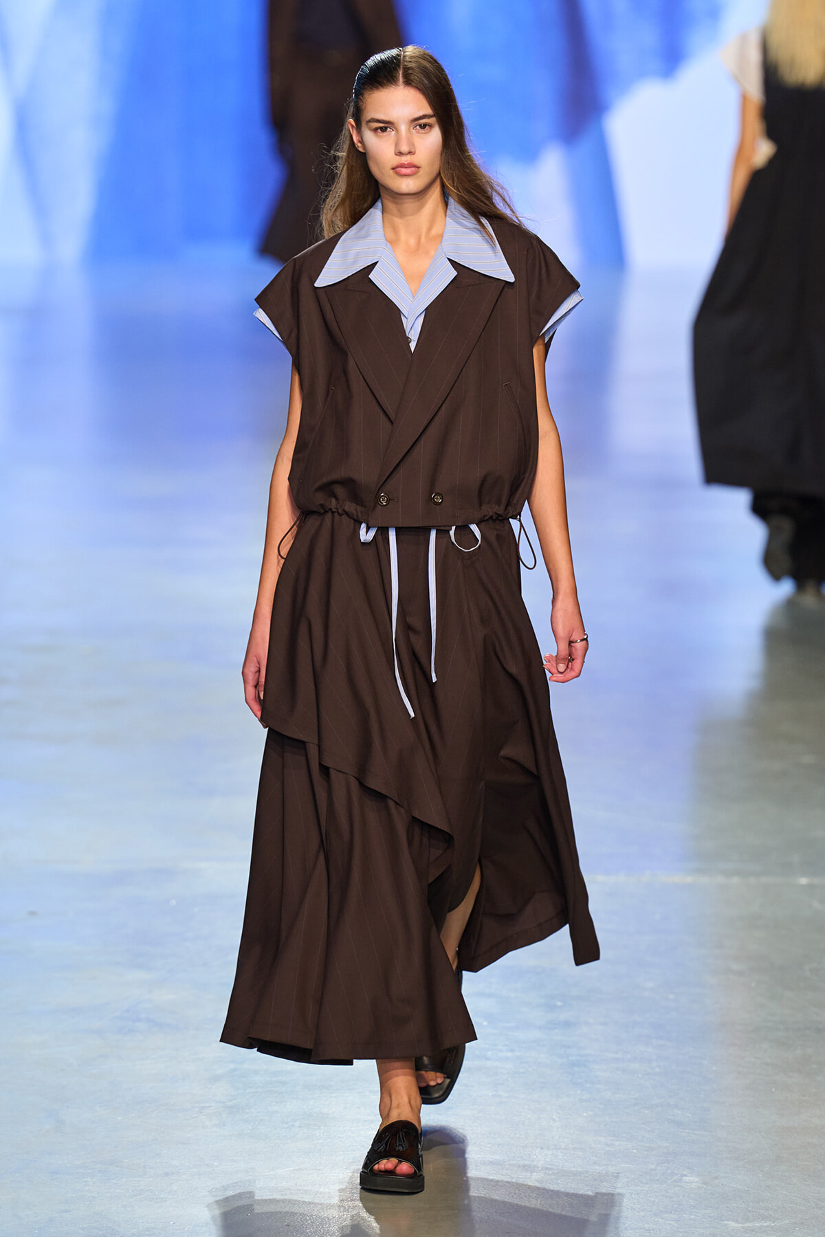 Model walking the runway in a brown pinstripe sleeveless dress with a large light-blue striped collar and a drawstring at the waist, paired with black sandals.