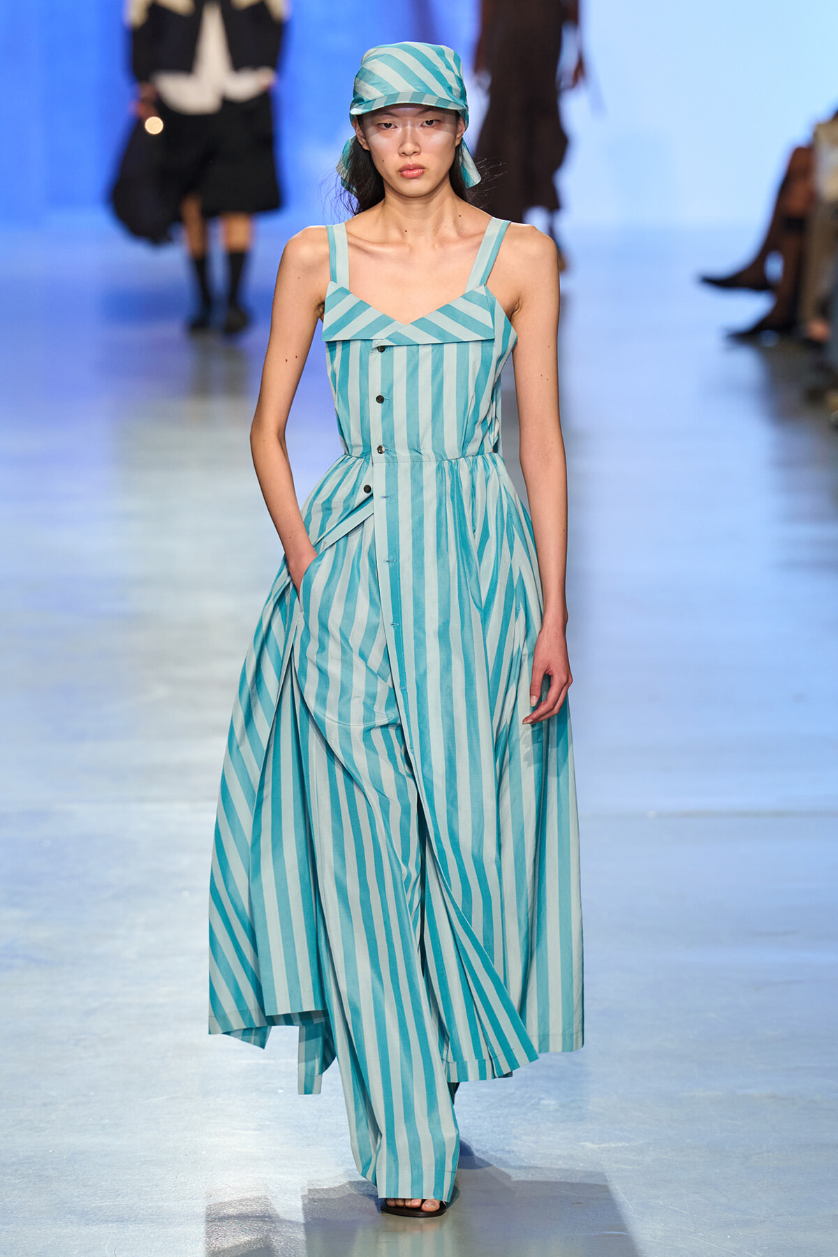 Model walking the runway in turquoise and white vertical-striped sleeveless jumpsuit with a matching headscarf and large front button details.