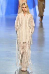 Model walks the runway wearing an ivory lace layered dress with fringe detailing and sheer panels.