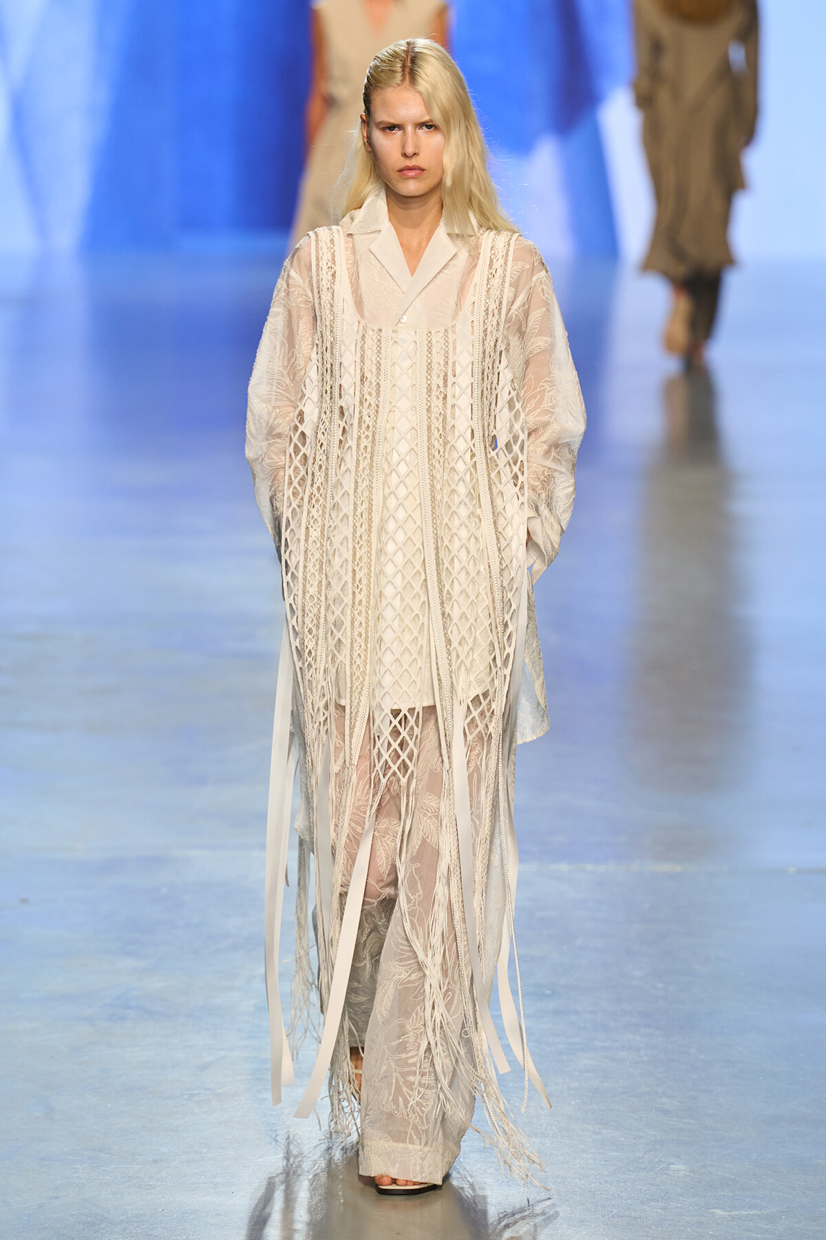 Model walks the runway wearing an ivory lace layered dress with fringe detailing and sheer panels.